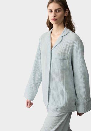 Vrouw draagt een losse, lichtblauwe pyjamaset met lange mouwen en een borstzakje, staand tegen een effen lichte achtergrond.