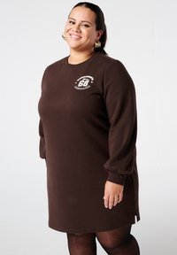 Bruine sweatjurk met lange, licht opgepofte mouwen. Heeft een ronde halslijn en een klein grafisch logo op de borst. Zachte, gestructureerde stof.