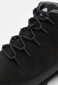 Gros plan d'une botte de randonnée noire avec des lacets épais et ronds et des œillets métalliques, montrant des détails cousus et un col de cheville rembourré.