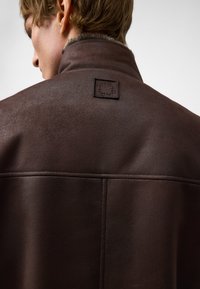 Bruine leren jas met een hoge kraag, voorzien van een gestructureerde afwerking en een logopatch op de rug. Strakke lijnen en een minimalistisch ontwerp.