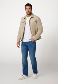 Šviesiai rudas zomšinis švarkas su dviem krūtinės kišenėmis, dėvimas virš baltos marškinėlių, derinamas su mėlynomis džinsomis ir baltomis sportbačiais. Paprastas, klasikinis dizainas.