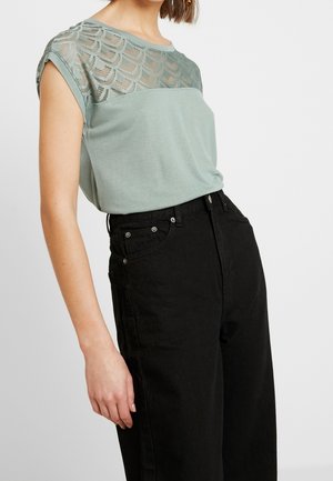 Kobieta w jasnozielonym krótkim topie z koronkowym wzorem na karczku i czarnych dżinsach z wysokim stanem, stojąca na białym tle.