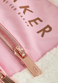 Ruusus kultainen vetoketjun vedin, jossa kirjain "B" vaaleanpunaisella kankaalla, jossa lukee "By Ted Baker", ja valkoista fleece-materiaalia.