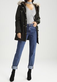 Femme portant un parka vert foncé à capuche en fourrure, un haut gris sans manches, un jean bleu retroussé et des bottines noires, debout devant un fond blanc.