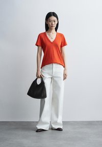 Γυναίκα με πορτοκαλί μπλουζάκι με V-λαιμό και λευκό πλατύ παντελόνι, κρατώντας μαύρη τσάντα, στέκεται σε γκρι πάτωμα με απλό γκρι φόντο.