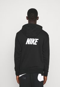 Hombre con sudadera negra de Nike y pantalones negros, de espaldas a la cámara frente a un fondo blanco liso.