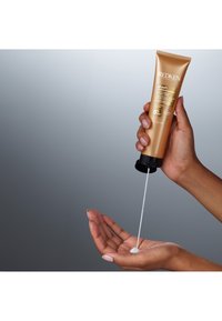 Redken All Soft Moisture Restore, złota tuba, czapka czarna, wydobywający białą kremową substancję na dłoń na szarym tle.