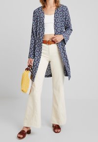 Blå mönstrad lång cardigan med öppen front, tillsammans med en vit cropped topp och krämfärgade utsvängda byxor. Brun bälte och vävd gul väska.