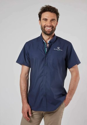 Chemise à manches courtes bleu marine avec un col intérieur rayé, arborant un logo sur la poche poitrine, fabriquée en tissu lisse.