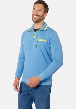 Lächelnder Mann mit kurzen Haaren und Bart trägt ein hellblaues Quarter-Zip-Sweatshirt mit gelben Reißverschlussakzenten und blaue Jeans, steht vor weißem Hintergrund.
