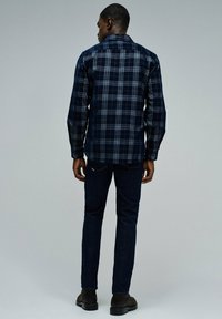 Camisa de cuadros azules con mangas largas, diseño de botones y corte recto. Combinada con jeans de mezclilla oscura y zapatos marrones.