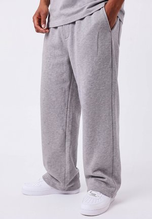 Partie inférieure du corps d'une personne portant un pantalon de survêtement gris ample et des baskets blanches, une main dans la poche, sur un fond uni.