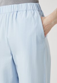 Svetlomodré nohavice vyrobené z hladkého materiálu, s elastickým pásom a bočnými vreckami, vystavené s rukou vo vrecku.