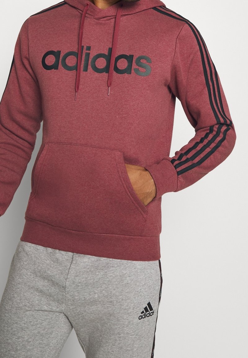 Personne portant un sweat à capuche Adidas bordeaux avec des bandes noires sur les manches et un pantalon de survêtement Adidas gris avec le logo sur la cuisse gauche.