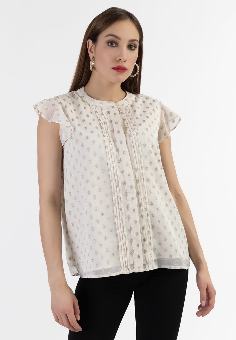 Camicia bianca con maniche corte a volant, caratterizzata da motivi floreali ricamati e pieghe verticali. Tessuto leggero e trasparente con una consistenza liscia.