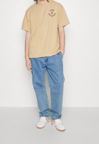 T-shirt en coton beige avec imprimé logo noir, associé à un jean en denim bleu clair et des baskets blanches avec des accents orange.