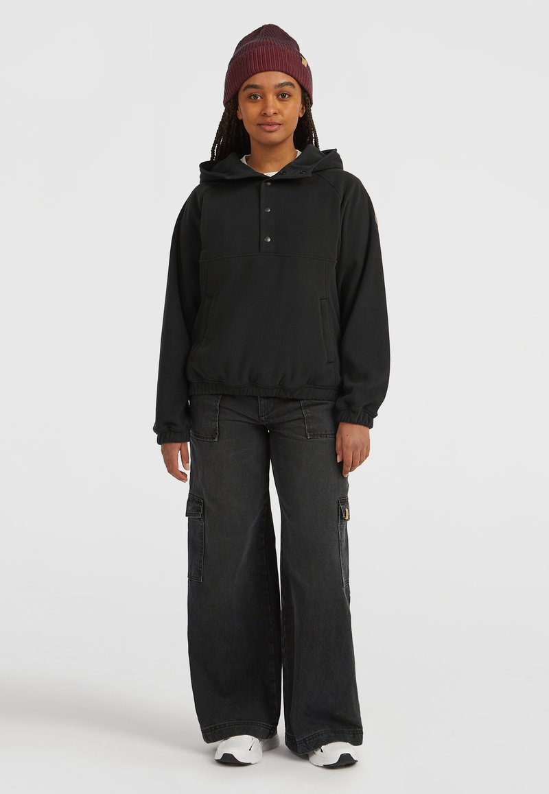 Schwarzer Kapuzenpullover mit Knopfleiste, kombiniert mit weit geschnittenen, dunklen Jeans-Cargohosen mit Seitentaschen. Weiße Sneakers und eine weinrote Mütze.