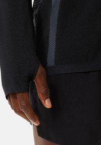 Zwarte sportieve top met lange mouwen en duimgaten, textuurstof, en een contrasterend blauw patroon langs de mouw en de zijnaad.