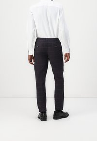 Geklede broek in marineblauw en bordeauxrood, met een elastische tailleband en twee achterzakken met knoopsluitingen. Zwarte schoenen maken de look compleet.