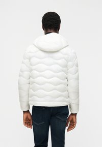 Άνδρας που φοράει λευ κό φουσκωτό μπουφάν με κουκούλα και σκούρο μπλε τζιν, στέκεται πλάτη σε απλό λευκό φόντο.