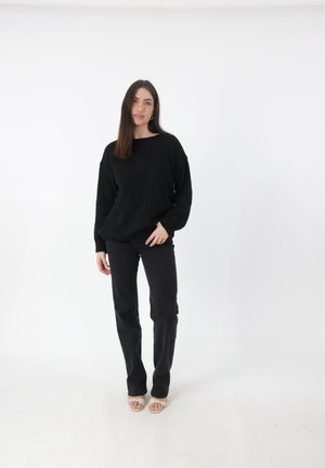 Zwarte, chunky gebreide trui met een brede halslijn en lange mouwen, gecombineerd met zwarte strakke jeans en neutrale open teenvandaag sandalen.