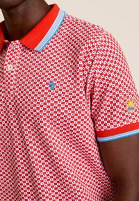 Rode polo shirt met korte mouwen, voorzien van een ruitpatroon in blauw en wit, met een contrasterende kraag en logo-opdruk op de mouw.