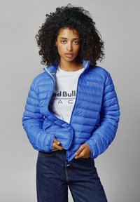 Plava zimska jakna s horizontalnim šavovima, visokim ovratnikom i žutim detaljima na zatvaraču. Nosena preko bijele majice s natpisom "Red Bull Racing".