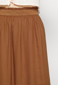 Donna che indossa una gonna marrone a vita alta con tessuto arricciato e una ampia cintura elastica, abbinata a un top floreale chiaro.