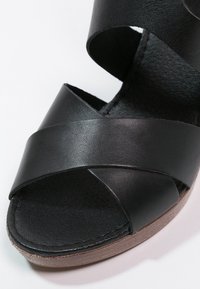Sandale en cuir noir avec des brides entrecroisées et une semelle intérieure noire texturée. Présente un bout arrondi et une semelle extérieure beige clair.