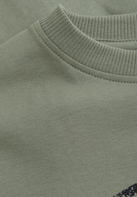 Grön bomull t-shirt tyg med ribbat runt halsringning. Slät textur med lätt stretch. Anmärkningsvärd kontrasterande grafisk detalj i mörk färg.