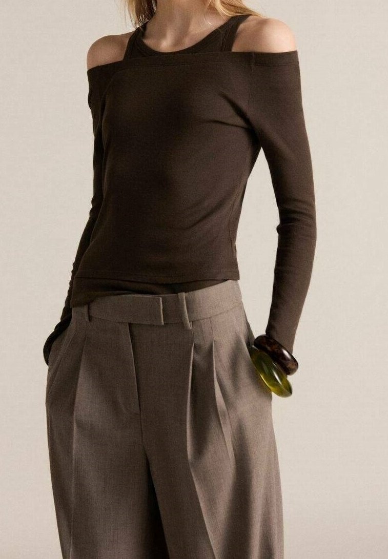 Haut brun foncé à épaules dénudées avec des manches longues, au design ajusté. Associé à un pantalon plissé gris clair, accessoirisé avec des bracelets en plastique.