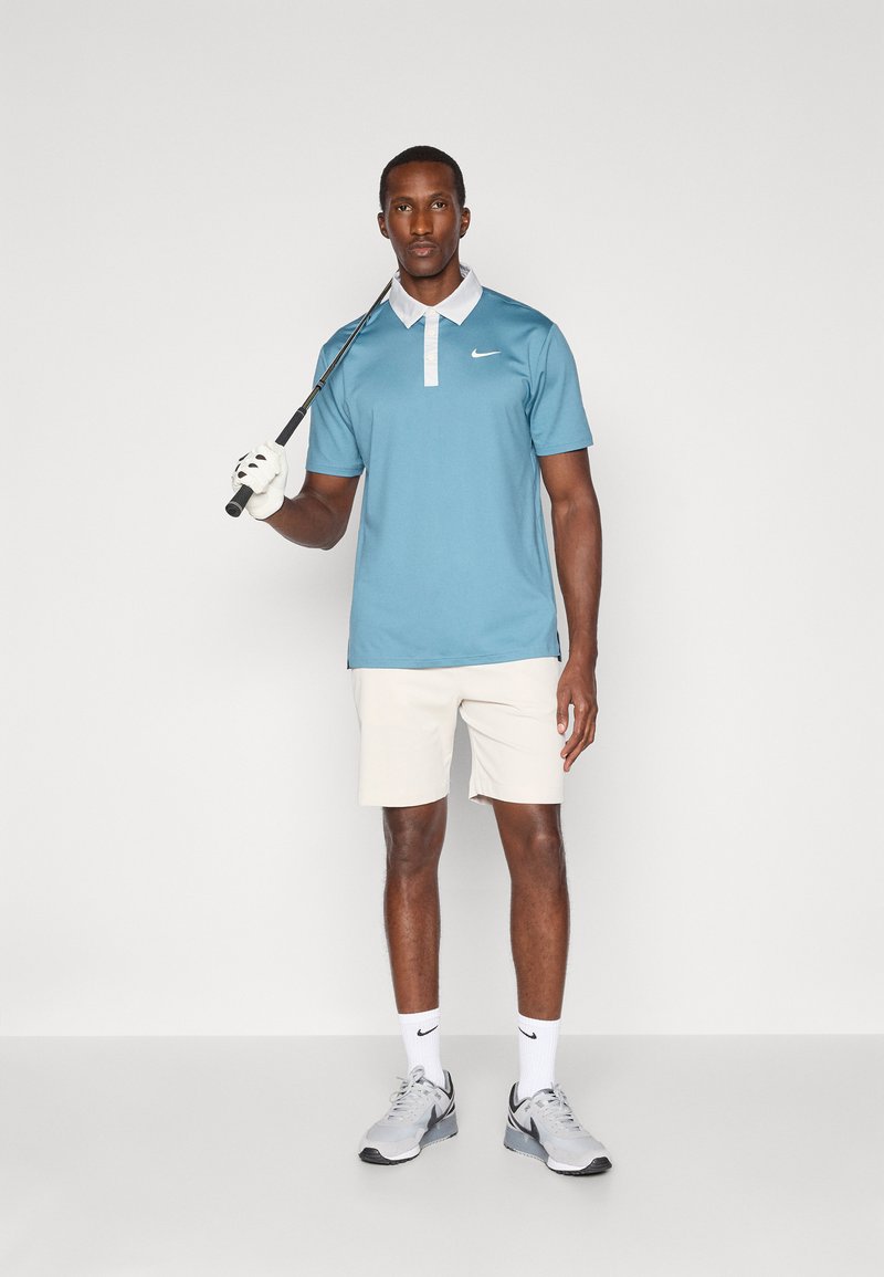 Lyseblå polo med hvid krave, beige shorts og grå Nike sneakers. Holder en golfkølle og har en hvid handske på.