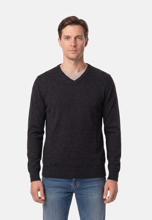 Uomo con maglione nero a V sopra una camicia beige e jeans blu, in piedi davanti a uno sfondo grigio uniforme.