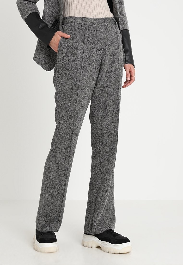 Person i grå texturerade byxor, beige ribbad tröja, grå kavaj med svarta läderärmar och svarta klumpiga sneakers med vita sulor.