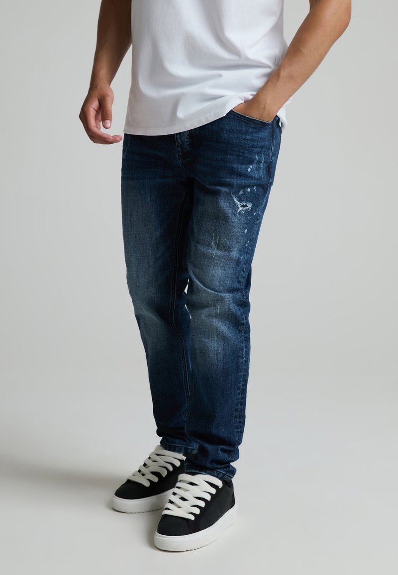 Homme portant un jean bleu foncé usé, des baskets noires avec des lacets blancs et un t-shirt blanc, une main dans la poche.