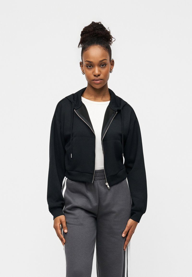 Μαύρο κοντό φούτερ με φερμουάρ και εμπρόσθιες τσέπες. Συνδυάζεται με γκρι φόρμα με λευκές ρίγες στο πλάι. Λείο υλικό.