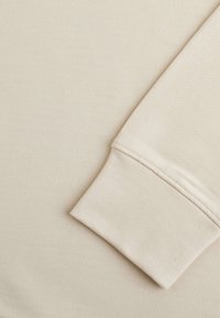 Tissu de sweat en beige avec une surface texturée. Un gros plan de la manche montre un bord doux et sans couture. Le matériau semble extensible et léger.