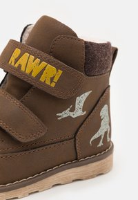 Bottines marron avec un accent en fourrure marron, un texte "RAWR!" jaune et des graphiques de dinosaure verts. Fermeture à velcro et semelle texturée.
