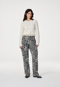 Crèmekleurige gebreide cardigan met gouden knopen, gecombineerd met hoge zwarte en witte panterprint broek. Model staat voor een effen achtergrond.