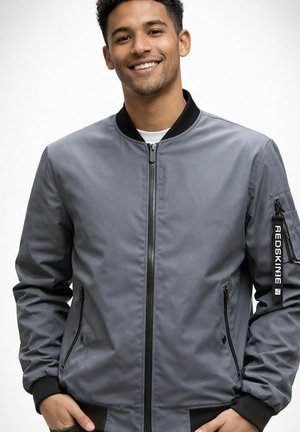 Homme souriant portant une veste bomber grise avec des poignets, un col et une fermeture éclair noirs, debout les mains dans les poches sur un fond blanc.