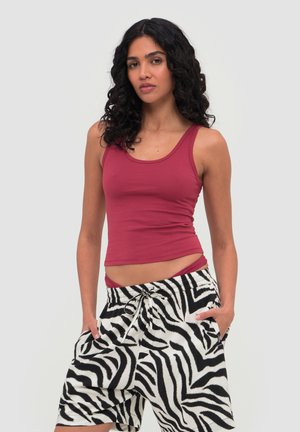Vrouw met krullend donker haar die een mouwloos bordeauxrood topje en zwart-witte zebra-print shorts draagt, met haar handen in haar zakken.