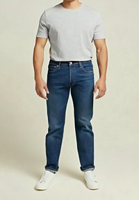 Homme debout en jean bleu foncé retroussé, baskets blanches décontractées et t-shirt gris clair à manches courtes sur un fond uni.