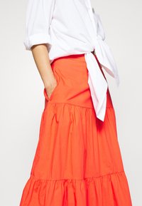 Henkilö, jolla on päällään valkoinen solmittu pusero, jonka hihat on kääritty, sekä korkeavyötäröinen, kerroksellinen, kirkkaan oranssi hame, ja yhdestä kädestä roikkuu käsi taskussa.