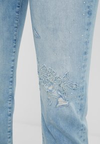 Jeans de mezclilla azul claro con detalles desgastados, bordado floral en el muslo y pequeños acentos de strass en toda la prenda.