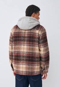 Chaqueta de franela a cuadros en marrón, crema y rojo con una capucha gris. Presenta un corte relajado y detalles de costuras visibles.