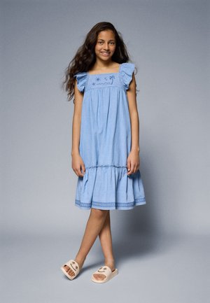 Light blue cotton dress with ruffled sleeves, featuring embroidered details on the chest and a tiered hem. Beige slide sandals with palm design.