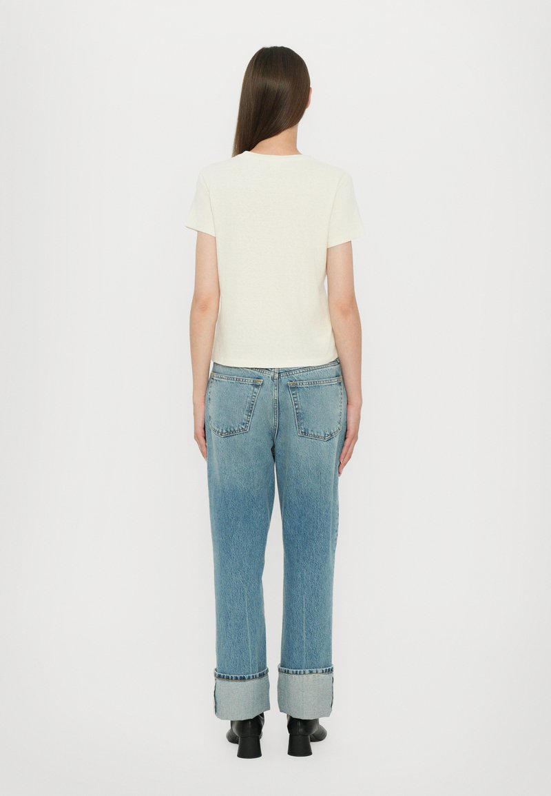 Cremfarbener T-Shirt mit kurzen Ärmeln, kombiniert mit hochgeschnittenen hellblauen Jeans mit umgeschlagenen Saum und Gesäßtaschen. Schwarze Knöchelschuhe.