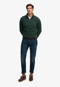 Ciemnozielony polar z półgolfem na suwak, zestawiony z niebieskimi dżinsami i brązowymi butami sznurowanymi. Codzienny krój o fakturowanej powierzchni.