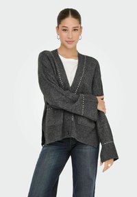Šedý pletený cardigan s ribbovaným lemem, knoflíkovým zapínáním a stříbrnými špičkami na přední části a rukávech. Nositelný přes bílý top a džíny.