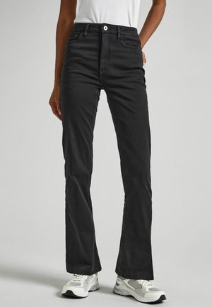 Osoba ubrana w czarne, wysokie spodnie z prostymi nogawkami, biały top na ramiączkach oraz białe trampki, stojąca na jednolitym tle.